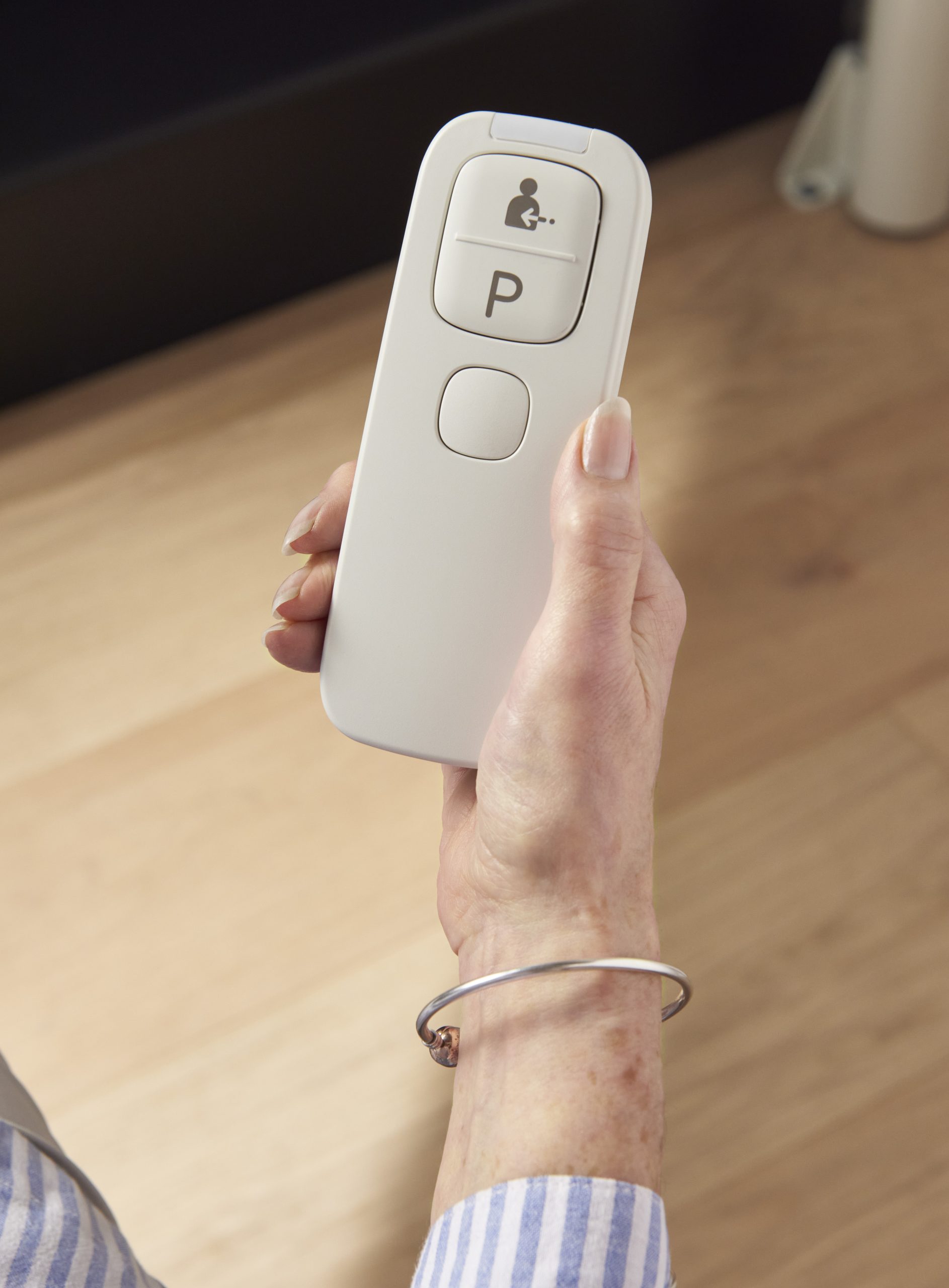 A close-up shot of an elderly person's hand holding a white, handheld remote control for a stairlift. The remote features large, simple buttons, including one with a "P" for parking and an icon depicting a person being called. The person is wearing a silver bracelet and a blue and white striped shirt, with a light oak floor visible in the blurred background.