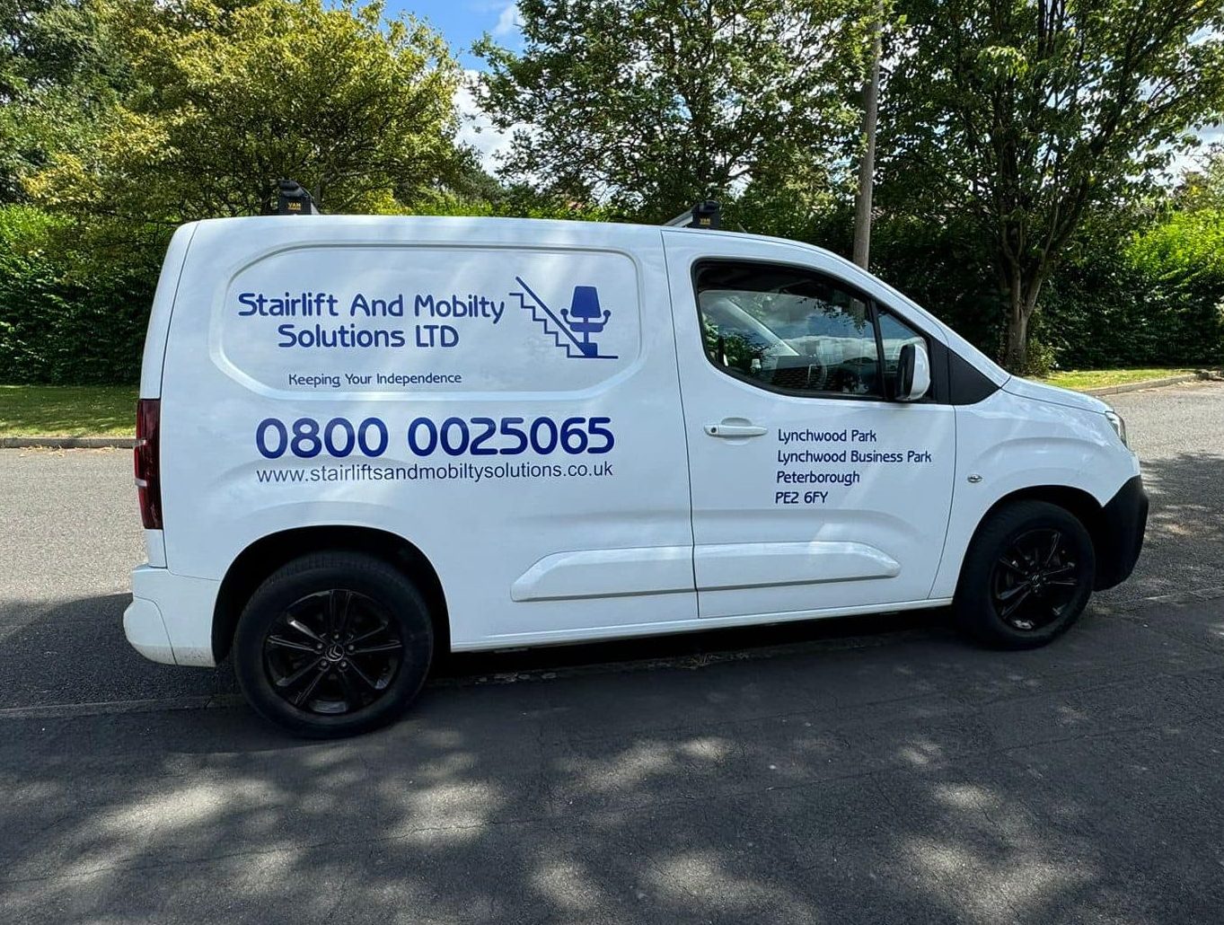 Gemini said A side-view of a white Stairlift And Mobility Solutions LTD service van parked on a residential street. The vehicle features blue branding with the company logo, 0800 contact number, and website on the main panel. The front door displays the local Peterborough business address at Lynchwood Business Park.
