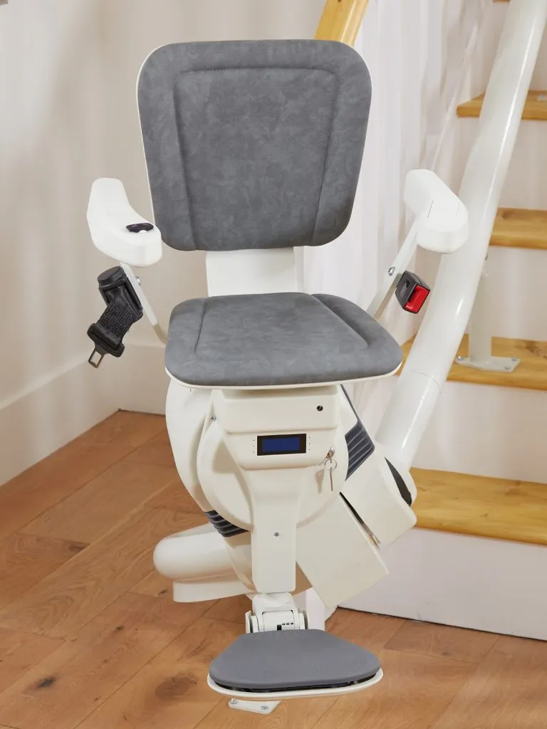 A front-facing view of a white curved stairlift with textured gray fabric upholstery on the seat, backrest, and footrest. The stairlift is mounted on a white tubular rail alongside a wooden staircase, featuring white armrests with integrated controls, a black seatbelt with a red buckle, and a small digital display screen below the seat with a key inserted in the ignition.