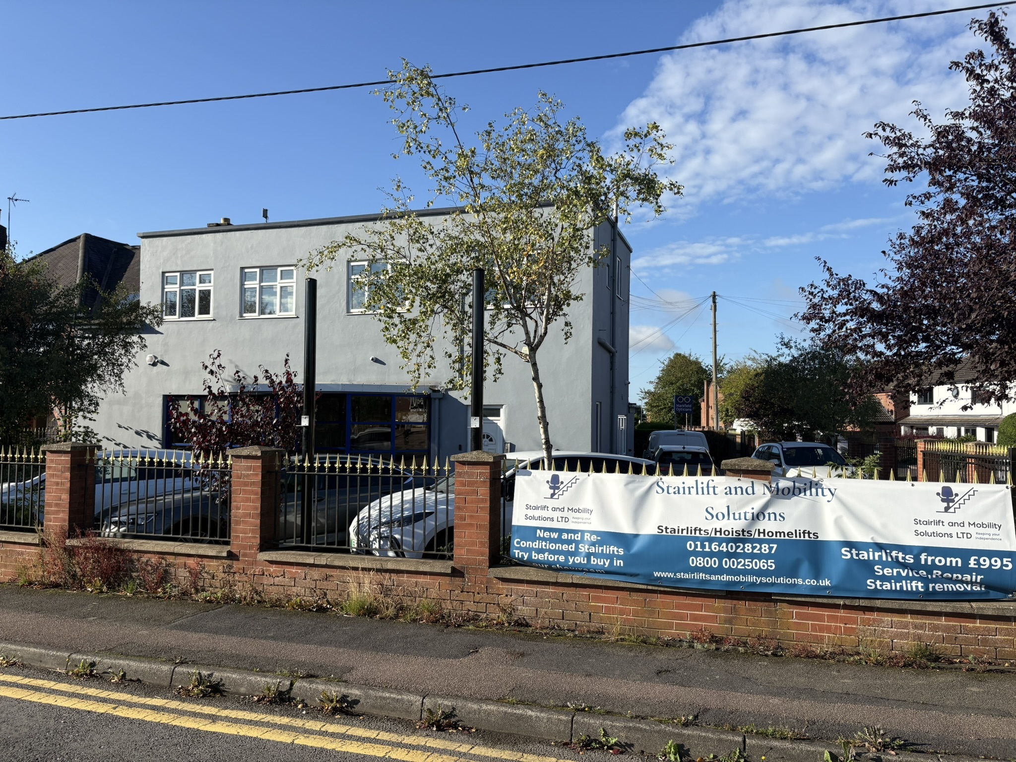 The exterior of the Stairlift and Mobility Solutions showroom, featuring a grey two-story building behind a brick wall and black metal railing. A large blue and white banner is displayed on the fence, advertising new and re-conditioned stairlifts, hoists, and homelifts, along with contact numbers and the company website.