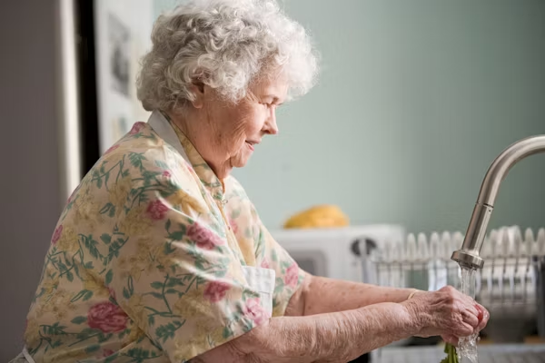An elderly woman with curly white hair, wearing a floral patterned shirt and a white apron, smiling gently while washing fresh green vegetables under a running kitchen tap.
