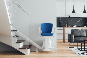 A modern, blue and white stairlift parked at the bottom of a contemporary staircase with wooden treads and a glass balustrade. The stairlift is positioned against a clean white wall in an open-plan living area, with a stylish kitchen and black marble backsplash visible in the background.