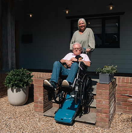 An elderly man in a manual wheelchair holding a small dog being transported down outdoor brick steps using a blue portable tracked stair climber, assisted by a smiling senior woman outside a house.